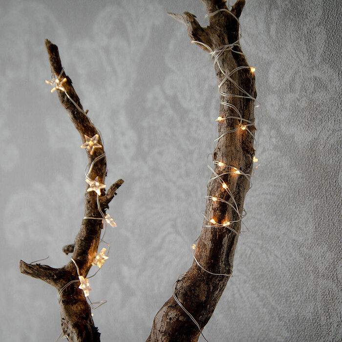Ein rustikaler Baumzweig ist detailreich mit einer filigranen Lichterkette umwickelt. Die kleinen, warmweißen LED-Lichter verleihen dem Ast eine festliche und zugleich gemütliche Stimmung. Der Hintergrund ist in weichen, neutralen Tönen gehalten, mit eine