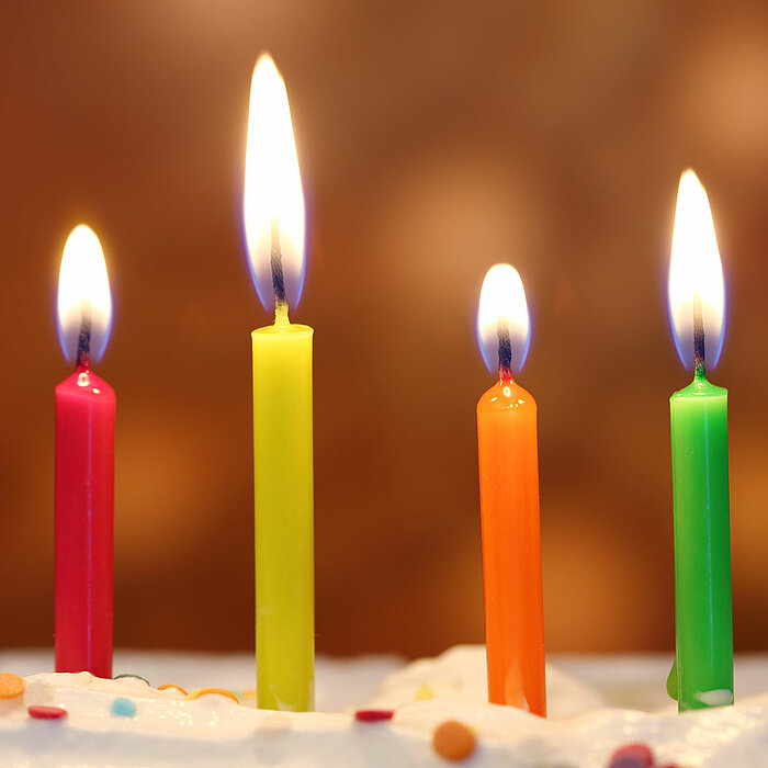 Vier brennende Geburtstagskerzen in leuchtenden Farben (Rot, Gelb, Orange und Grün) stecken in einem weißen Kuchen mit bunten Zuckerstreuseln. Der unscharfe Hintergrund in warmen Tönen verleiht dem Bild eine festliche und freudige Stimmung.