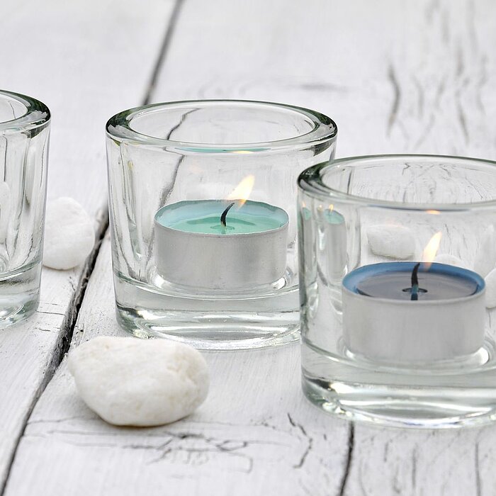 Drei brennende Kerzen von Müller, zwei in zylindrischen Betonhaltern und eine in einem runden Glas, umgeben von Streublumen und Blütenblättern auf einer weißen Oberfläche. Dazwischen liegen zwei kleine, herzförmige Schokoladen. Im Bild sind auch zarte wei