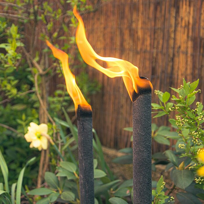 Zwei brennende Bolsius Kerzen in geflochtenen Kerzenhaltern stehen auf einem gelben, filigran geformten Tisch im Garten. Im Hintergrund befindet sich eine Holzbank mit hellgrünen, beigen und pinken Kissen sowie Brennholz darunter. Auf der Bank liegen ein 