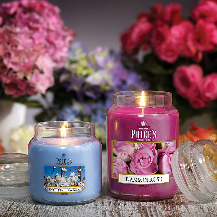 Drei brennende kerzenpinker "Prices Candles" in verschiedenen Größen stehen auf einem goldenen Tablett, umgeben von violetten und weißen Blüten. Links davon befindet sich Raumduft von "Prices Candles" mit mehreren Holzstäbchen in einer Flasche. Rechts ist