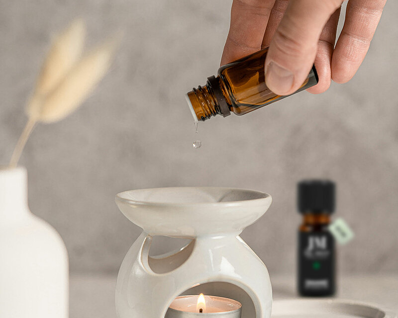 in der Mitte des Bildes sieht man ein weißes Duftstövchen auf einer ovalen betonschale. In dem Stövchen steht ein sanft brennendes Teelicht. Man sieht wie ein Mann ein kleines Fläschchen mit Aroma Öl auf den oberen Teller des Stövchens schüttet. Im Hintergrund steht ein weiteres Fläschchen mit Ätherischem Öl und eine weiße Vase mit einem getrockneten Farnzweig. Das gesamte Bild ist hell freundlich und harmonisch.