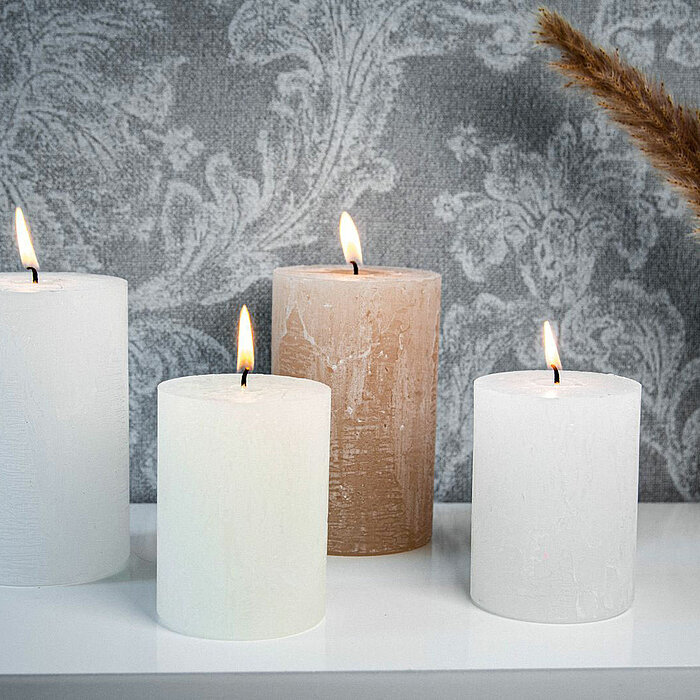 Vier brennende Bolsius Rustikkerzen in Weiß und Beige stehen auf einem weißen Regal vor einer grauen, ornamental gemusterten Tapete. Rechts neben den Kerzen steht eine durchsichtige Vase mit getrockneten Gräsern.