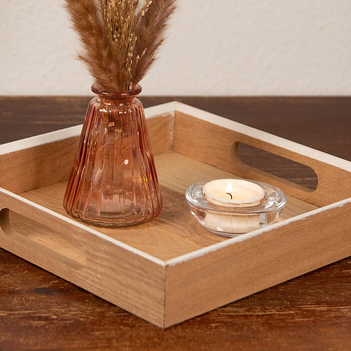 Ein hölzernes Tablett steht auf einem Holztisch. Auf dem Tablett befindet sich eine kleine Vase aus rosa getöntem Glas, die mit trockenen Gräsern gefüllt ist. Neben der Vase steht ein kleiner Glas-Kerzenhalter mit einem Teelicht. Die Szene strahlt eine ge