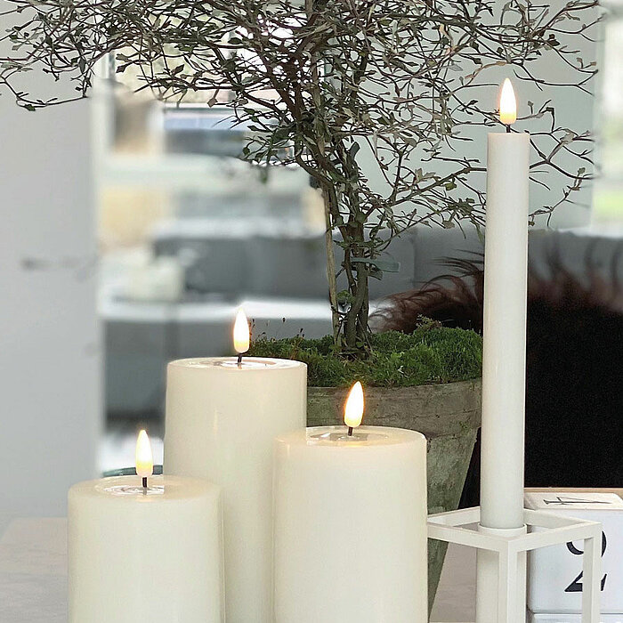 Vier brennende weiße Stumpenkerzen vom Hersteller "Deluxe Homearts" stehen auf einer spiegelnden Oberfläche. Drei der Kerzen sind in verschiedenen Größen und die vierte ist eine schlanke, lange Kerze in einem modernen Kerzenhalter. Dahinter befindet sich 