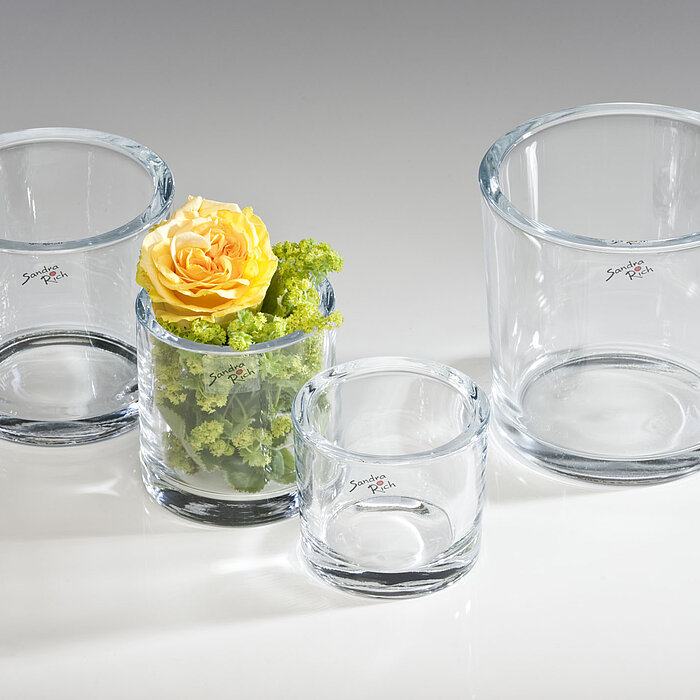 Drei leere Glasvasen vom Hersteller "Sandra Rich" in verschiedenen Größen stehen auf einer hellen Oberfläche. Eine der Vasen ist mit einer gelben Rose und grünen Blüten dekoriert.