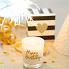 Eine einzelne, brennende Kerze in einem Glas mit der Aufschrift "Happy Birthday" steht in der Mitte einer Feierdekoration. Über der Kerze sprüht ein Wunderkerze helle Funken. Im Hintergrund sind goldene Bänder, ein gestreiftes Geschenk mit einem goldenen 