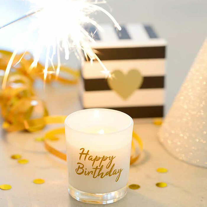 Eine einzelne, brennende Kerze in einem Glas mit der Aufschrift "Happy Birthday" steht in der Mitte einer Feierdekoration. Über der Kerze sprüht ein Wunderkerze helle Funken. Im Hintergrund sind goldene Bänder, ein gestreiftes Geschenk mit einem goldenen 