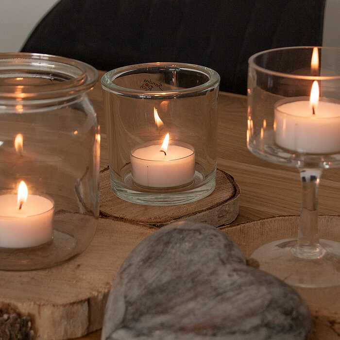 Drei brennende weiße Teelichter, die in verschiedenen Glasgefäßen stehen. Das erste Gefäß ist ein großes Einmachglas, das zweite ein kleineres Glas mit einem breiten Rand, und das dritte steht in einem eleganten Glas auf einem Stiel. Die Gläser stehen auf
