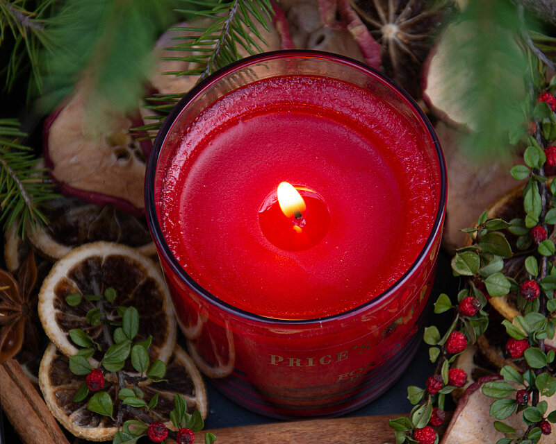 Eine mit armen Kerzenlicht brennende Duftkerze im Glas mit dem Duft Apple-Spice von Prices steht in der Mitte eines festlich dekorierten Tisches. Umgeben von verschiedenen Tannenzweigen, Beeren, getrockneten Orangenscheiben und Zimtstangen verbreitet sie einen gemütlichen und einladenden Duft.