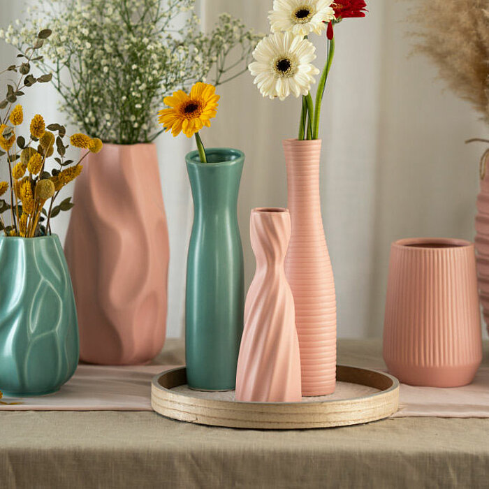 Drei Vasen vom Hersteller "Sandra Rich" in den Farben Creme und Dunkelbraun stehen auf einem runden Tisch mit einem Drahtgestell. Im Hintergrund sind ein Kamin mit gestapeltem Brennholz und ein graues Sofa mit einem gemusterten Kissen zu sehen. Die cremef