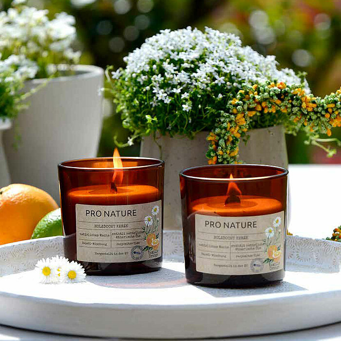 Das Bild zeigt zwei brennende Kerzen in Glasgefäßen des Herstellers Müller, die die Aufschrift "Pro Nature" tragen. Die Kerzen stehen auf einem Holztisch, umgeben von lila und pinkfarbenen Blumen sowie einigen grünen Blättern. Im Hintergrund ist eine drit
