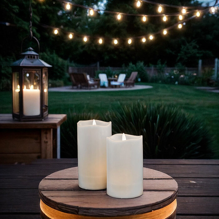 Zwei brennende weiße Stumpenkerzen stehen auf einem hölzernen Tablett, das auf einem dunklen Holztisch platziert ist. Im Hintergrund befindet sich ein Garten mit leuchtenden Lichterketten, die eine gemütliche Atmosphäre erzeugen. Links im Bild steht eine 