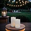 Zwei brennende weiße Stumpenkerzen stehen auf einem hölzernen Tablett, das auf einem dunklen Holztisch platziert ist. Im Hintergrund befindet sich ein Garten mit leuchtenden Lichterketten, die eine gemütliche Atmosphäre erzeugen. Links im Bild steht eine 