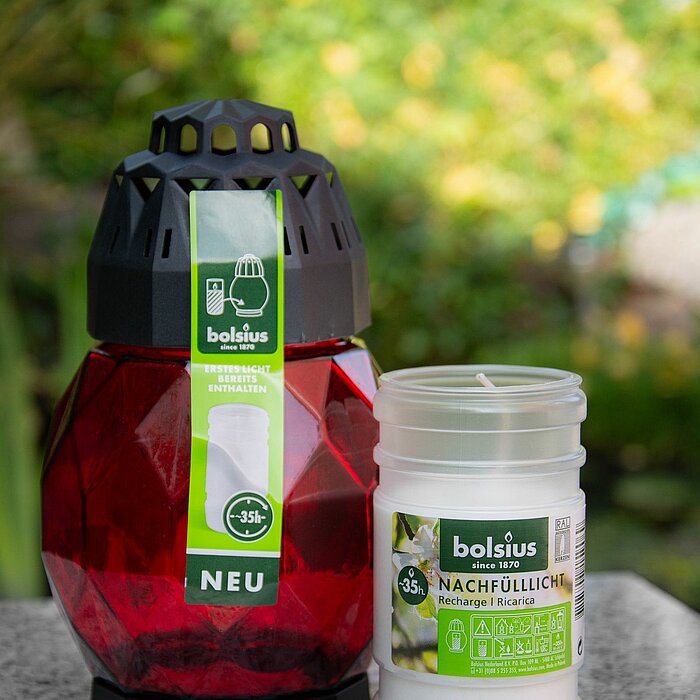 Eine rote Bolsius Grablaterne mit einem schwarzen Deckel, daneben eine weiße Bolsius Nachfüllkerze für die Grablaterne, beide auf einem grauen Tisch im Freien. Der Hintergrund zeigt einen Garten mit grünen Pflanzen und verschwommenen Blumen, was eine ruhi