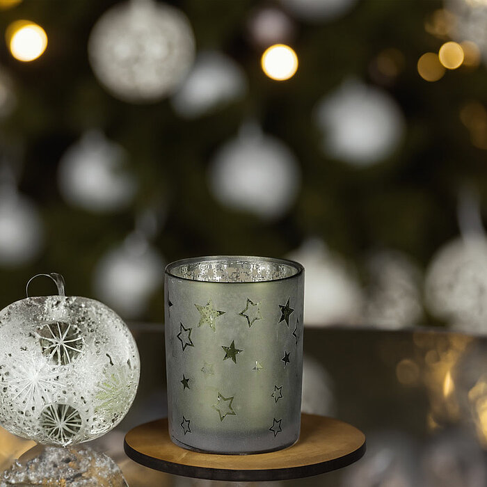 Ein Edelman Teelichthalter mit Sternenmustern steht auf einem Holztisch. Im Hintergrund ist ein weihnachtlich geschmückter Baum zu sehen, sowie Geschenkbänder und dekorative Folien in Gold und Weiß. Daneben liegen kunstvoll gefaltete Papierblumen in Rot u