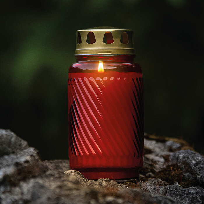 Eine einzelne leuchtende Bolsius Grabkerze mit rot-geriffelten Glas steht auf einem grauen, moosbewachsenen Stein. Der goldene Deckel der Kerze hat Löcher für Luftzirkulation. Der Hintergrund ist dunkel und verschwommen, was die Kerze im Vordergrund beson