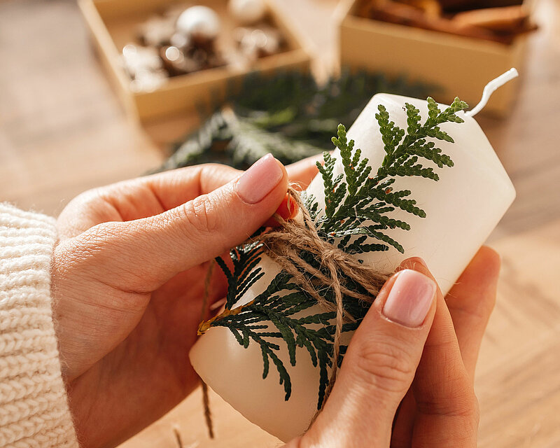 Auf einem Tisch stehen mehrere Bastelmaterialien. Man sieht wie eine Frau eine Stumpenkerze in der Hand hält und mit einer Jutekordel ein Fichtenzweig an der Kerze anbringt.