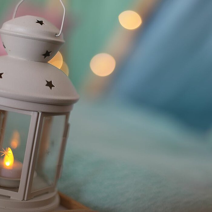 Eine kleine weiße Laterne mit einem brennenden Teelicht darin steht auf einer weichen, türkisfarbenen Decke. Die Laterne hat sternförmige Aussparungen an den Seiten. Im Hintergrund sind verschwommen bunte Lichter und eine gemütliche, farbenreiche Atmosphä