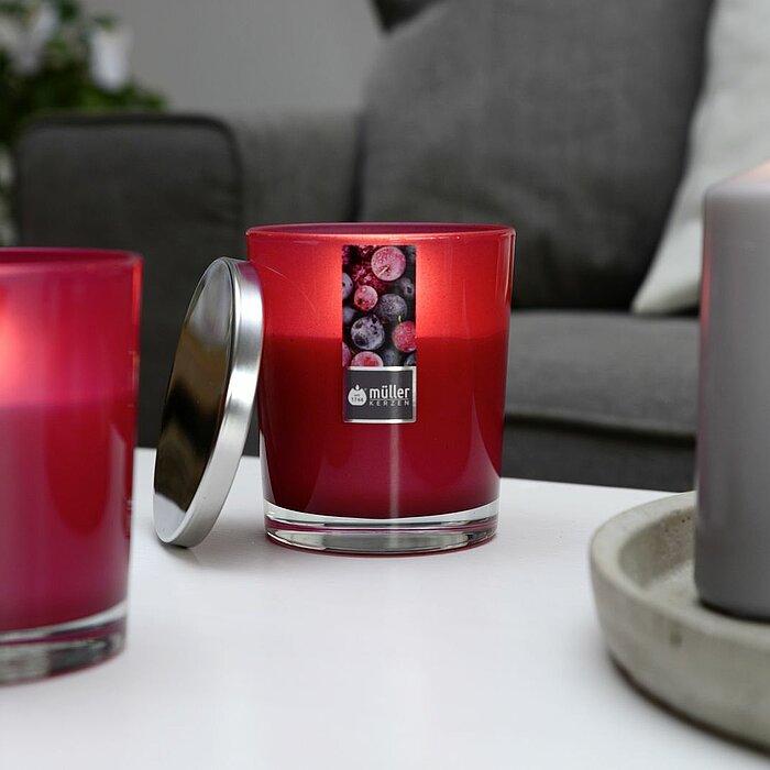 Fünf brennende Kerzen des Herstellers Müller, platziert auf einer runden, grauen Schale. Die Kerzen variieren in der Größe und Form, darunter zwei Glasbehälter-Kerzen, eine Stumpenkerze und zwei Teelichter. Im Hintergrund befindet sich ein graues Sofa mit