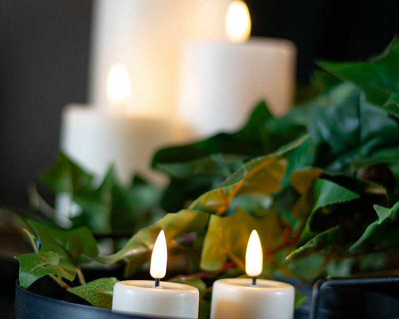 Auf einem schwarzen Dekoteller stehen im Vordergrund zwei sanft flackernde, weiße LED-Teelichter von Deluxe Homeart. Der Teller ist mit künstlichem Efeu dekoriert, was dem Arrangement eine natürliche Note verleiht. Im Hintergrund erkennt man drei unterschiedlich hohe, ebenfalls weiße LED-Stumpenkerzen von Deluxe Homeart, die sanft flackern und dem Arrangement eine ruhige, entspannende Ausstrahlung verleihen.