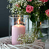 Eine brennende rosa Müller-Kerze in einem Glaswindlicht, platziert in einem weißen geflochtenen Korb. Im Hintergrund steht ein Strauß aus rosa Rosen und weißen Schleierkraut-Blüten in einer klaren Vase auf einem Tisch. Die Kombination verleiht dem Bild ei