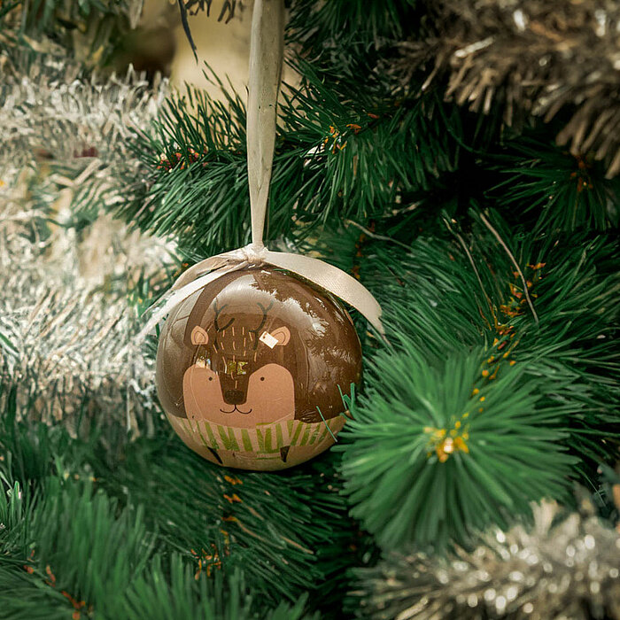 Eine weihnachtliche Szene zeigt eine Nahaufnahme eines Tannenbaums. An einem der Zweige hängt eine Christbaumkugel mit einem Schneemann-Motiv und einer kleinen weißen Schleife. Die Kugel ist detailreich gestaltet und strahlt eine festliche Atmosphäre aus.