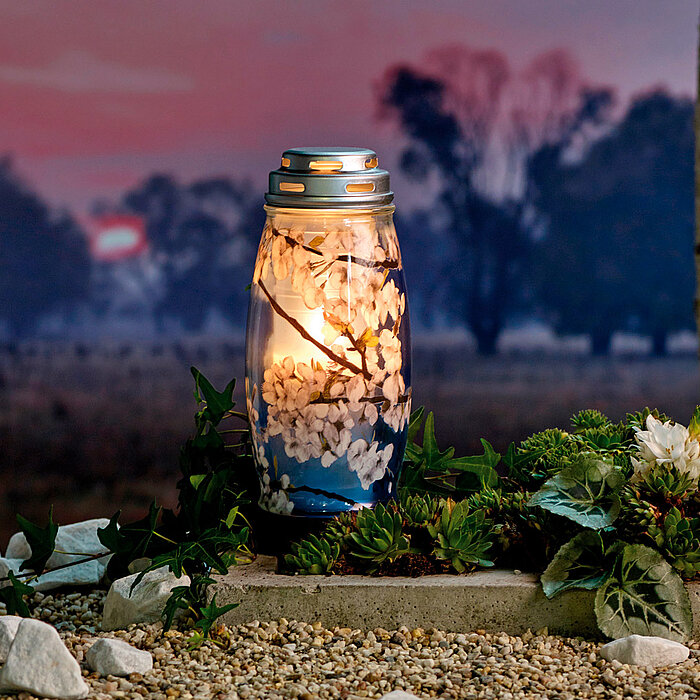 Eine Laterne, die mit einer brennenden Kerze erleuchtet ist, steht auf einem Kiesbett, umgeben von Pflanzen und weißen Steinen. Im Hintergrund erstreckt sich eine Landschaft mit Bäumen vor einem malerischen, farbenfrohen Sonnenuntergang.