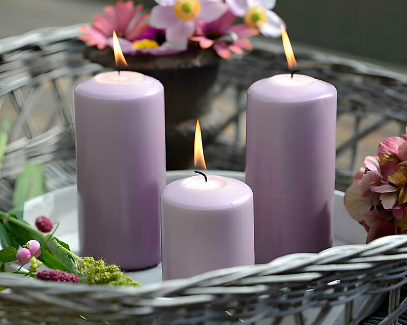 Drei brennende Stearin-Stumpenkerzen in zartem Flieder, stehen in einem Flechtkörbchen zwischen verschiedenen Blüten in Rosa und Weiß.