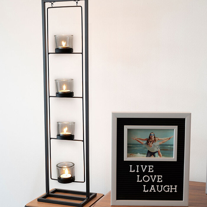 Vier brennende Teelichtkerzen in Glasbehältern befinden sich in einem hohen, schwarzen Metallkerzenhalter, der auf einem Tisch steht. Daneben steht ein gerahmtes Bild mit der Aufschrift "LIVE LOVE LAUGH" und einem Foto von zwei Personen, die am Strand pos