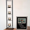 Vier brennende Teelichtkerzen in Glasbehältern befinden sich in einem hohen, schwarzen Metallkerzenhalter, der auf einem Tisch steht. Daneben steht ein gerahmtes Bild mit der Aufschrift "LIVE LOVE LAUGH" und einem Foto von zwei Personen, die am Strand pos