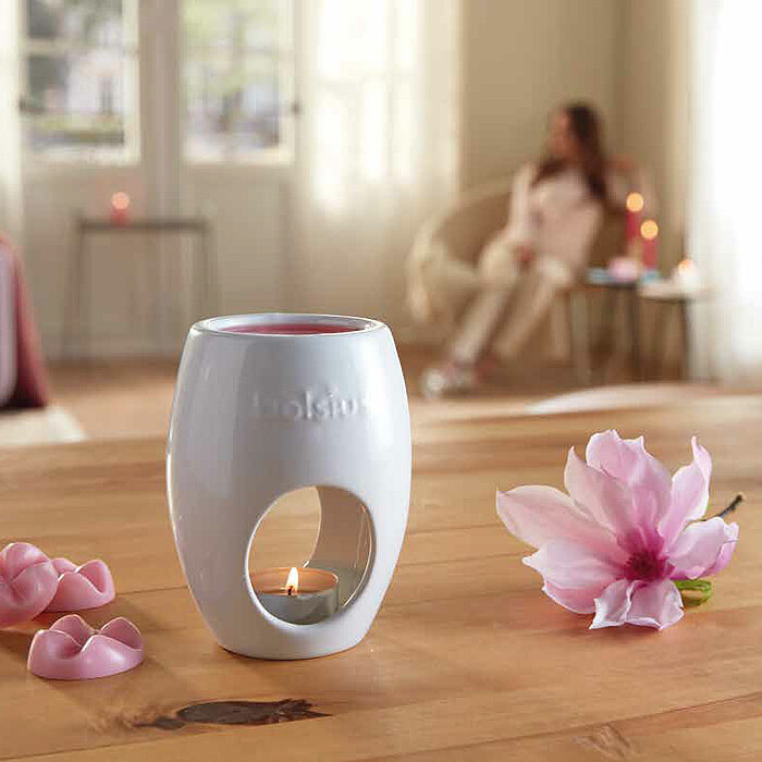 Eine Frau stellt eine rosa Duftkerze von Bolsius in einen weißen Kerzenhalter auf einem Holztisch. Auf dem Tisch befinden sich weitere rosa Bolsius-Duftkerzen in unterschiedlichen Formen, eine Glasvase mit rosa Blumen und eine Teetasse. Im Hintergrund ist