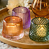 Auf dem Bild sind drei brennende Teelichter in Glasbehältern vom Hersteller "Sandra Rich" zu sehen. Die Glasbehälter haben unterschiedliche Designs in den Farben Lila, Orange und Grün. Sie stehen auf einem runden Tablett aus geflochtenem Holz. Im Hintergr