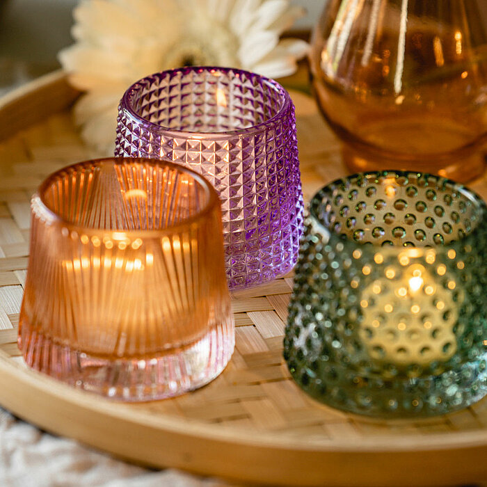 Auf dem Bild sind drei brennende Teelichter in Glasbehältern vom Hersteller "Sandra Rich" zu sehen. Die Glasbehälter haben unterschiedliche Designs in den Farben Lila, Orange und Grün. Sie stehen auf einem runden Tablett aus geflochtenem Holz. Im Hintergr