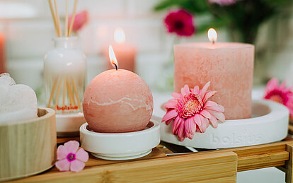 Eine brennende Rustik-Kugelkerze in zartem Rosa steht neben einer Rustik Stumpenkerze auf weißen Untersetzern zwischen rosa Blüten.