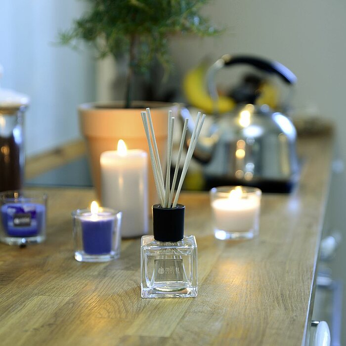 Vier brennende Müller Kerzen in verschiedenen Farben und Formen stehen auf einem hölzernen Küchentresen. Zwei der Kerzen sind in durchsichtigen Gläsern, eine dicke weiße Stumpenkerze und eine kleine violette Kerze. Neben ihnen steht ein Müller Raumduft mi