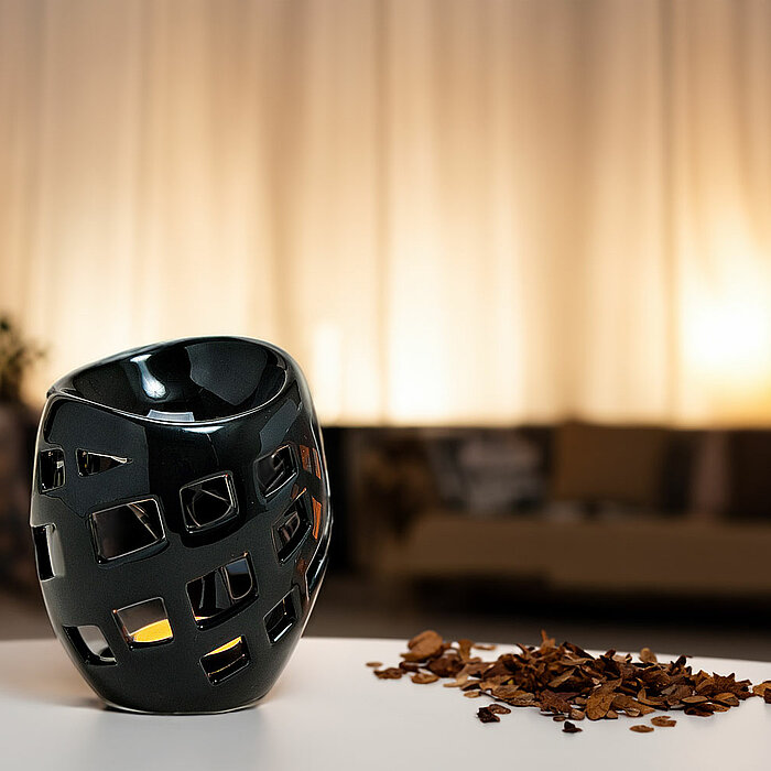 Auf einem weißen Tisch steht ein dunkler Kerzenhalter mit vielen Aussparungen. Im Hintergrund brennt schwaches Licht, und es sind verschwommene Konturen eines gemütlichen Wohnzimmers mit einem Sofa zu erkennen. Auf dem Tisch liegen neben dem Kerzenhalter 