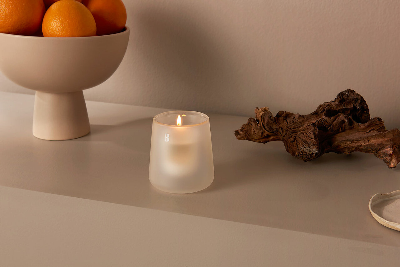 Ein milchweißes Kerzenglas mit umweltfreundlichem Nachfüller steht auf einer modernen Kommode neben einem Stück Treibholz und einer Schale mit Orangen.
