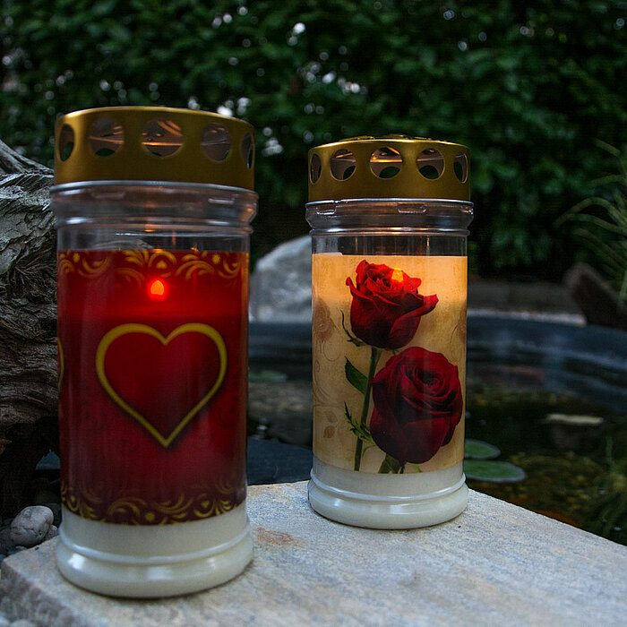 Zwei Grabkerzen mit goldenem Deckel auf einem Stein vor einem Teich. Die linke Kerze ist rot mit einem Herzen als Motiv, die rechte Kerze zeigt zwei rote Rosen auf hellem Hintergrund. Sie stehen auf einem Stein, umgeben von kleinen Steinen und einem Stück