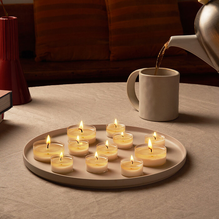 Acht brennende Bolsius Teelichter stehen auf einem weißen Tablett, das auf einem runden Tisch platziert ist. Daneben liegt ein Stapel Bücher und eine rote Vase. Eine Hand gießt Tee aus einer silbernen Kanne in eine weiße Tasse. Die Szene vermittelt eine g