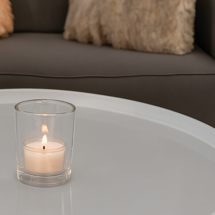 Zwei brennende weiße Teelichter von DecoLite in transparenten Glashaltern stehen auf einer weißen, marmorierten Unterlage. Rechts davon liegt eine einzelne, kräftig pinke Gerbera-Blume. Die Szene wirkt ruhig und harmonisch.