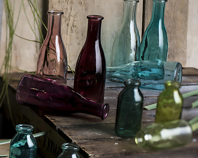 Das gesamte Bild hat eine rustikale und angenehme Ausstrahlung. auf verschieden hohen Holzboxen stehen und liegen unterschiedlich farbige Deko-Flaschen von Sandra Rich.