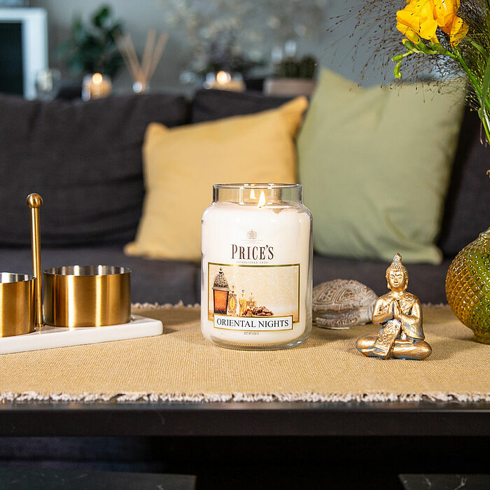 Eine brennende Kerze von "Price's Candles" in einem Glas mit der Aufschrift "WINTER JASMINE" steht auf einem weißen Regal. Daneben liegt ein Tannenzapfen und einige grüne Zweige. Links von der Kerze befindet sich eine türkisfarbene Vase mit getrockneten B