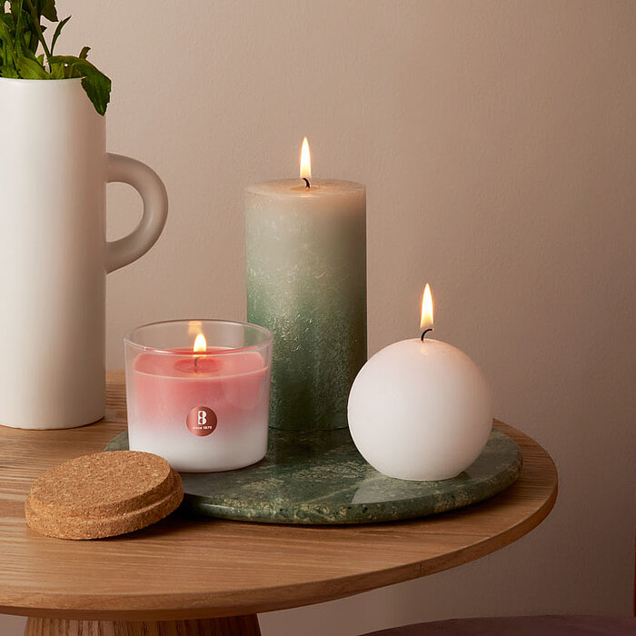 Eine brennende Bolsius Rustikkerze in Zylinderform mit einem verzierten Muster in Rosa sowie eine runde Bolsius Rustikkerze in Rosa stehen auf einem runden, rosafarbenen Tablett auf einem Holztisch. Daneben befinden sich ein rotes Bolsius Duftkerzenglas, 