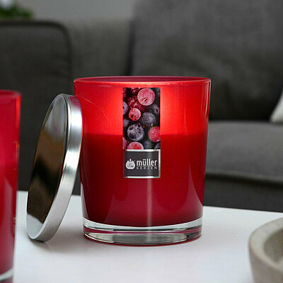 Eine Nahaufnahme von einem weißen Wohnzimmertisch vor einem grauen Sofa. Auf dem Tisch steht eine brennende, graue Stumpenkerze auf einem Betonteller. Links daneben stehen zwei brennende Duftkerze in einem dunkelroten Glas und einem silbernen Deckel daneben.