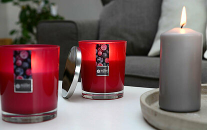 Eine Nahaufnahme von einem weißen Wohnzimmertisch vor einem grauen Sofa. Auf dem Tisch steht eine brennende, graue Stumpenkerze auf einem Betonteller. Links daneben stehen zwei brennende Duftkerze in einem dunkelroten Glas und einem silbernen Deckel daneben.