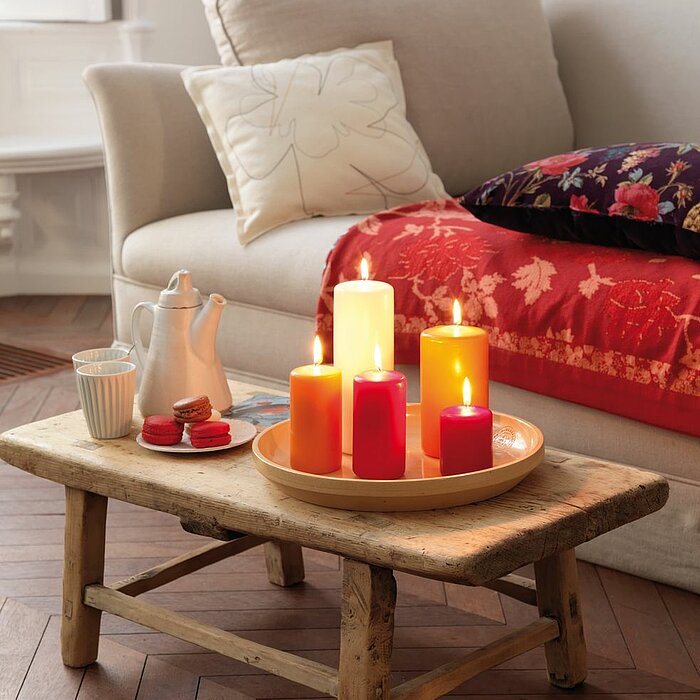 Neun brennende Bolsius Stumpenkerzen in verschiedenen Größen und Farben (Lila, Rot, Pink, Gelb und Grün) stehen auf einem runden silberfarbenen Tablett, das auf einem kleinen Holztisch im Wohnzimmer platziert ist. Im Hintergrund erkennt man ein beigefarbe