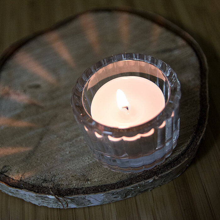Drei brennende Teelichter des Herstellers Sandra Rich in klaren Glasbehältern. Zwei davon sind zylindrisch und befinden sich auf einer Holzplatte, während das Dritte in einem herzförmigen Glashalter auf einem hölzernen Tisch steht. Die Umgebung ist minima