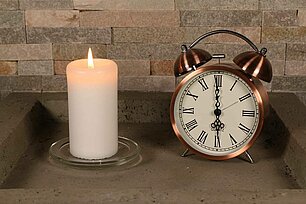 In einer eckigen grauen Betonschale steht eine brennende weisse Bolsius professional Stumpenkerze mit den Maßen 120/58 mm. Rechts neben der Stumpenkerze befindet sich ein schöner alter Wecker in Kupferfarbe, der die Uhrzeit 6 Uhr anzeigt. Im Hintergrund sieht man eine steinerne Mauer.