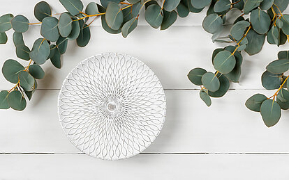 Das gesamte Bild strahlt ruhe aus und ist in hellen Tönen gehalten. Auf einem weissen Holzboden steht ein weisser Dekoteller mit einem grünen gestreiften Muster. Oberhalb am Bildrand sind einige Blätterzweige in sanftem grün.